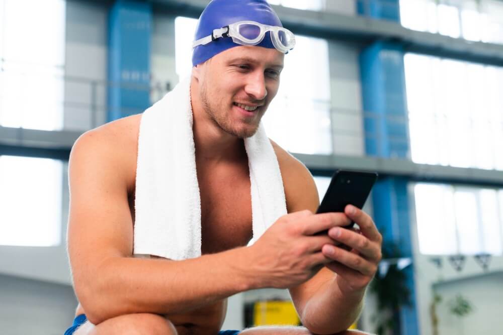 a fit adult man relaxing at an indoor swimming facility after exercise. He is wearing a swim cap and goggles, with a towel around his neck, and is using a smartphone while seated beside the pool.