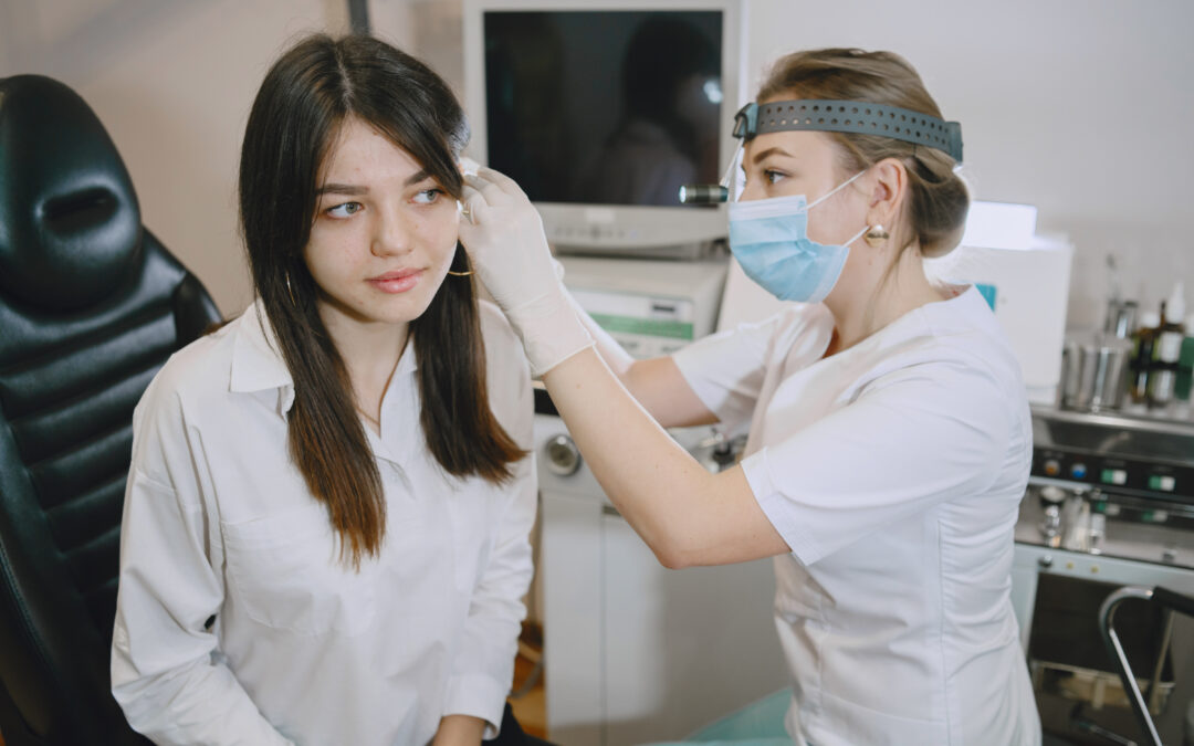 a female healthcare professional wearing protective equipment examining a young female patient’s ear with a handheld otoscopic instrument.